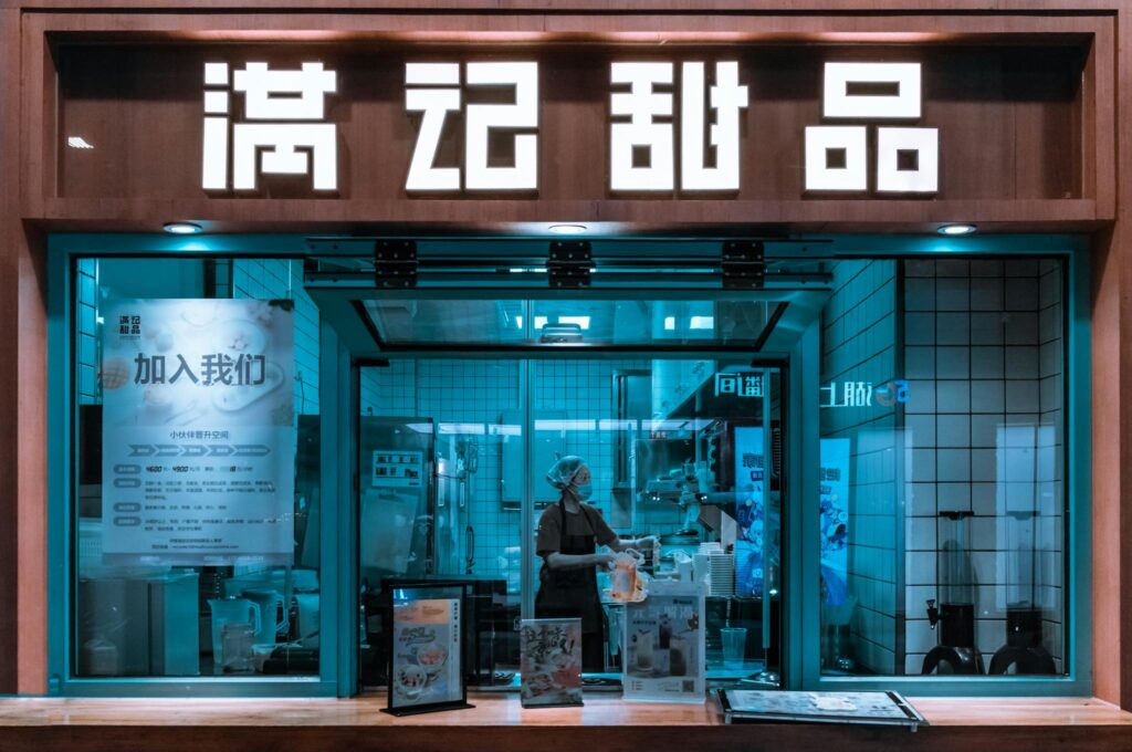 A modern restaurant kitchen viewed from outside at night, featuring a worker inside.